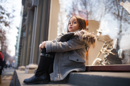 Child girl in a coat with a fur hood is sitting next to shopwindow on city street in evening. Closeup.の写真素材