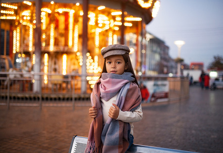 Child girl in a cap and scarf is standing at the street against background of evening city lights.の写真素材