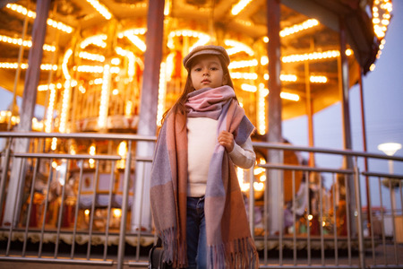 Child girl in a cap and scarf is standing at the street against background of carousel and evening city lights.の写真素材
