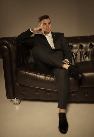 Young businessman in a black suit and a white shirt is sitting and pondering on the leather sofa against a brown background.の写真素材