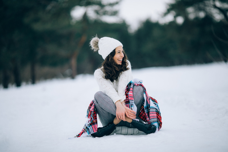 Beautiful woman is sitting and laughing on the snow dressed in white sweater, hat and scarf during the winter walk.の写真素材