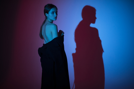 Young naked woman stands in an unbuttoned coat and casts her shadow on the wall. Backlight. Back view.の写真素材