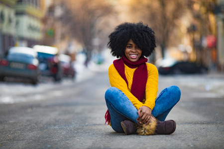 Cheerful young black woman in scarf and sweater is sitting in the middle of the street.の写真素材