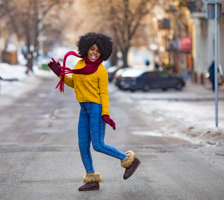 Cheerful young black woman in scarf and sweater is walking to the city street and smiling.の写真素材
