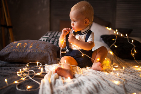 Baby boy sits on the bed and plays with garland of glowing light bulbs.の写真素材