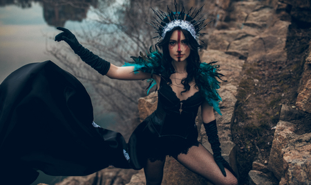A young woman in the image of a fairy and a sorceress standing next to rocks over a lake in a black dress and a crown.の写真素材