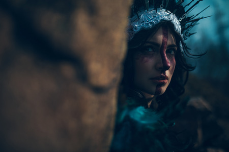 Portrait of young woman in the image of a fairy and a sorceress standing next to the rock in a black dress and a crown.