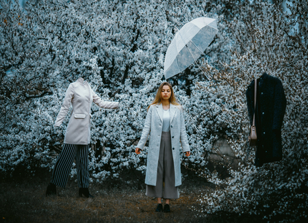 A young woman stands in a blooming garden among mannequins and an umbrella flying in the air. The...