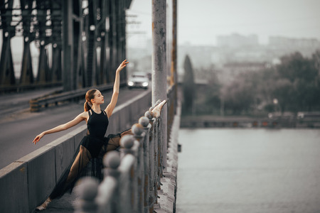 Ballerina sitting in twine pose in a black transparent dress on the bridge through the river against the background of cityscape.の写真素材