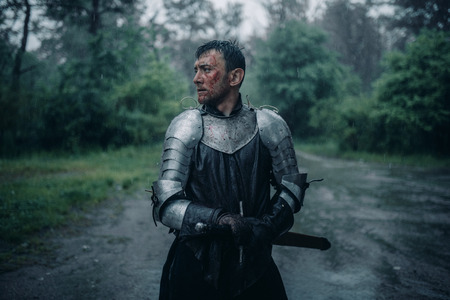 A young man stands in the image of a medieval knight in armor and with a sword in his hands in the rain.の写真素材