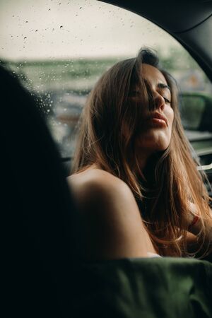 A young woman sits inside a car with bare shoulders against the background of raindrops on the...