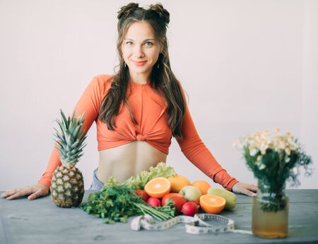 A young smiling woman is standing next to vegetables, fruits and a measuring tape on a table. The concept of diet and healthy eating.の写真素材