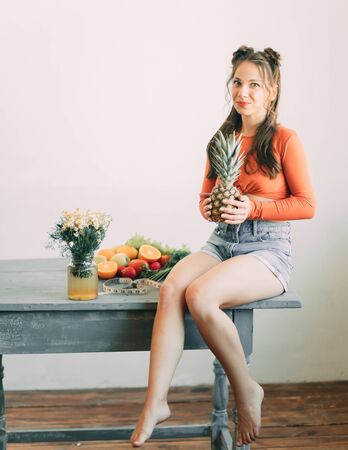 A young smiling woman is sitting with pineapple in her hands next to vegetables, fruits and a measuring tape on a table. The concept of diet and healthy eating. Place for copy space.の写真素材