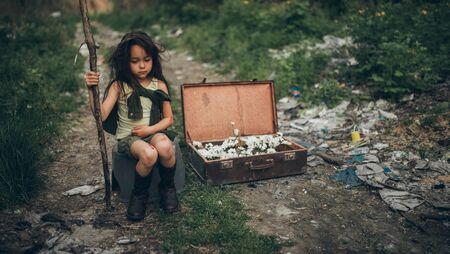 A homeless girl is sitting on a garbage dump next to a suitcase with flowers inside. The concept of environmental pollution and landfill reclamation. The concept of poverty and homelessness.の写真素材