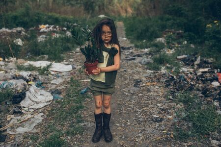 A homeless girl is standing on a garbage dump with a houseplant in a pot. The concept of...