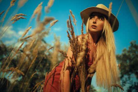 Young blond woman in a hat is on a meadow among grass ears.