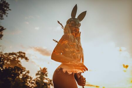 Young blond woman stands topless in black mask of bunny on meadow. The effect of double multiple exposure.の写真素材