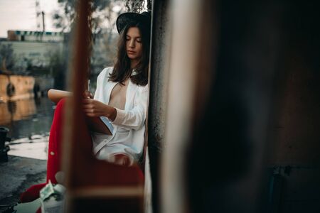 A young woman sits and draws in sketchbook on an old abandoned ship in an unbuttoned white shirt, hat and red tights.の写真素材