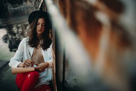 A young woman sits and draws in sketchbook on an old abandoned ship in an unbuttoned white shirt, hat and red holey tights.の写真素材