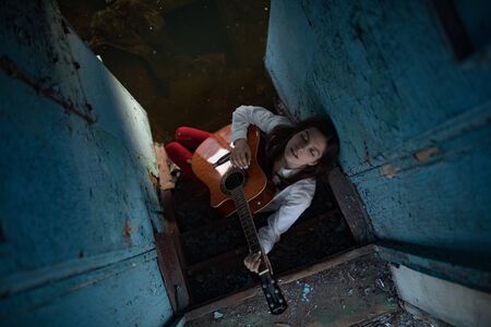 A young woman sits and plays guitar inside of old abandoned ship. Top view.