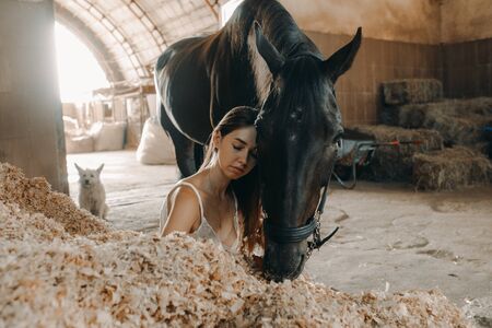 A young woman sits on a pile of wood shavings in a stable next to a standing horse.の写真素材