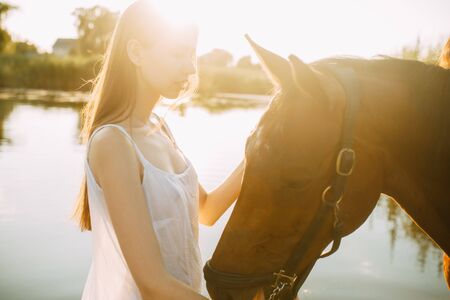 A young woman stands next to horse on background of river at sunset. Backlight.の写真素材