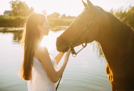 A young woman stands next to horse on background of river at sunset. Backlight.の写真素材