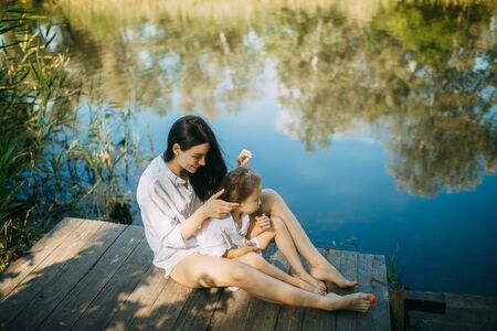 Mother and daughter are sitting on a wooden bridge over a river with reflection of trees and sky in the water.の写真素材
