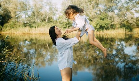 Mother and daughter play and have fun over a small river with reflection of trees and sky in the water.の写真素材
