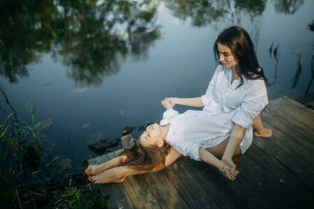 Mother and daughter play and have fun on a wooden bridge on background of river.の写真素材
