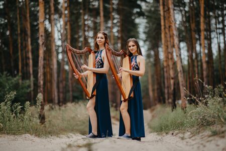Two women harpists stand at forest and play harps in beautiful dresses against a background of pines.の写真素材