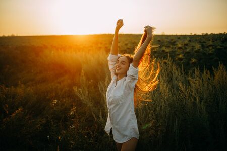 Happy joyful woman in white shirt is walking with pleasure among the flowers and grass on meadow at sunset.の写真素材
