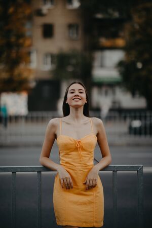A young woman stands on the sidewalk in a yellow dress on a background of cityscape.の写真素材
