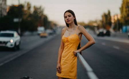 A young woman stands on the sidewalk in a yellow dress on a background of cityscape.