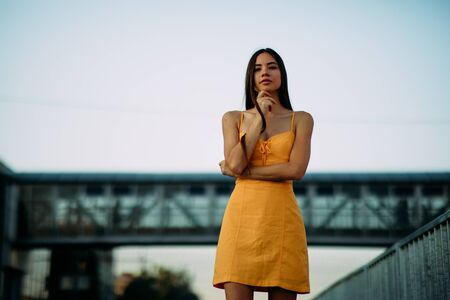 A young woman stands in a yellow dress on a background of a bridge and sky.の写真素材