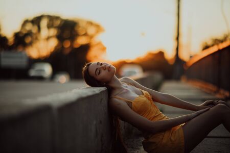 A young woman sits on the sidewalk in a yellow dress on a background of cityscape at sunset.の写真素材