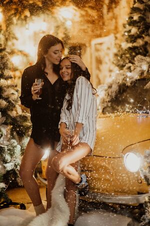 Lesbian couple holds glasses of wine in their hands and stands against the background of Christmas decorations, snowfall and a yellow retro car.の写真素材