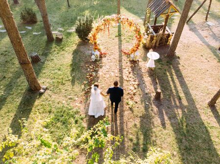 The bride and groom hold hands and go to the wedding arch in a pine forest. Drone shooting.