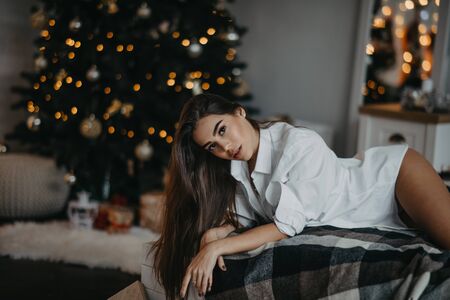 Young woman in unbuttoned white shirt lies on the bed against the background of the Christmas tree and decorations.の写真素材