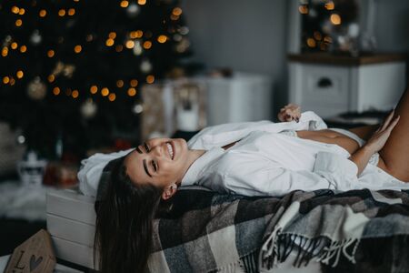 Young woman in unbuttoned white shirt lies on the bed and smiles against the background of the Christmas tree and decorations.の写真素材