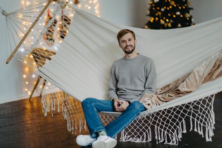Happy young man sits in hammock against the background of the Christmas tree and garlands.の写真素材