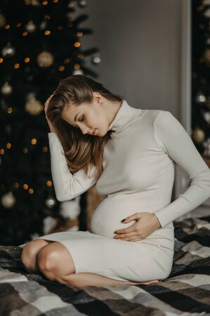 Young pregnant woman in white dress sits on the bed against background of Christmas tree.の写真素材