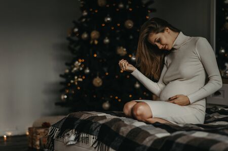 Young pregnant woman in white dress sits on the bed against background of Christmas tree.の写真素材