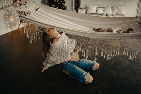 Young pregnant woman in pullover and jeans sits at the floor next to hammock against background of Christmas tree and decorations.の写真素材
