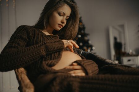 Young pregnant woman sits in the armchair and touches her tummy by her hand.の写真素材