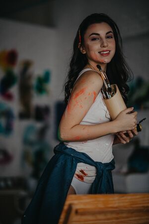 The artist stained with paint stands in an art workshop and holds box with paintbrushes in her hands.の写真素材