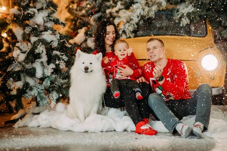 Happy family with little son sits next to white samoyed dog against background of Christmas tree, retro car and snowfall.の写真素材