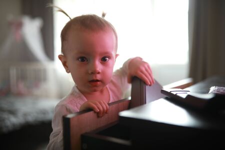 Toddler girl stands next to the table at the room against the window.
