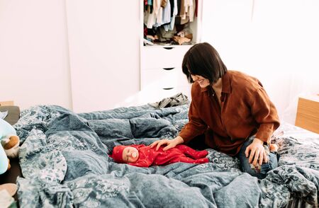 Mother sits at the bedroom next to a lying baby on the bed.の写真素材