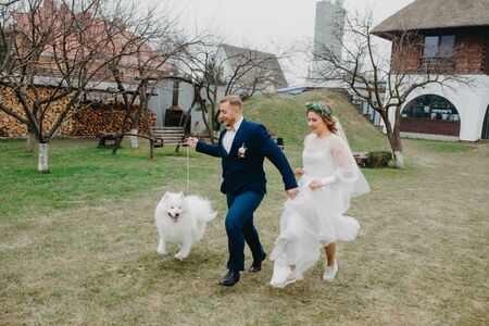 Newlyweds have a fun and run on the lawn next to the Samoyed dog on the background of the house.の写真素材